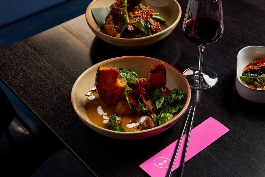 Bowl of pumpkin curry resting on a dark wooden table with a glass of red wine.