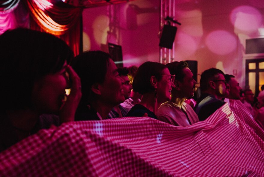 A dark image of people in a row holding up a striped sheet with deep pink lighting on the whole room. 