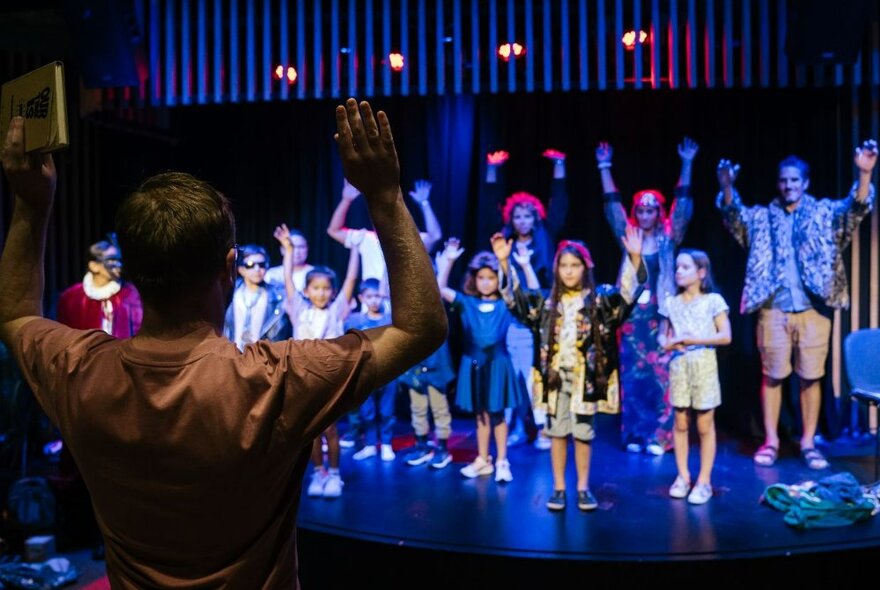 A group of children and adults standing on a stage, with their arms raised in the air above them.