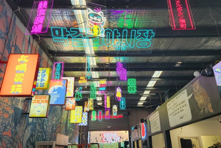 Looking up into the ceiling of a large spacious hall showings flags, neon food signs, banner signage and lanterns.