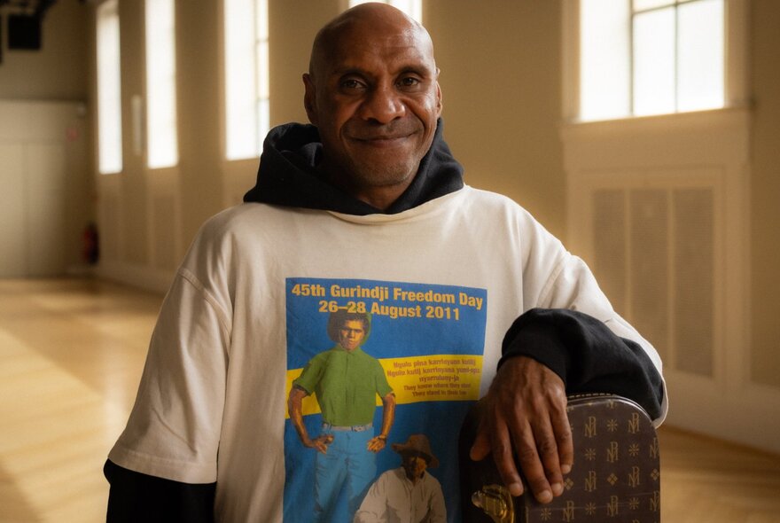 A smiling Amos Roach, wearing a white slogan t-shirt and standing in a large empty room.