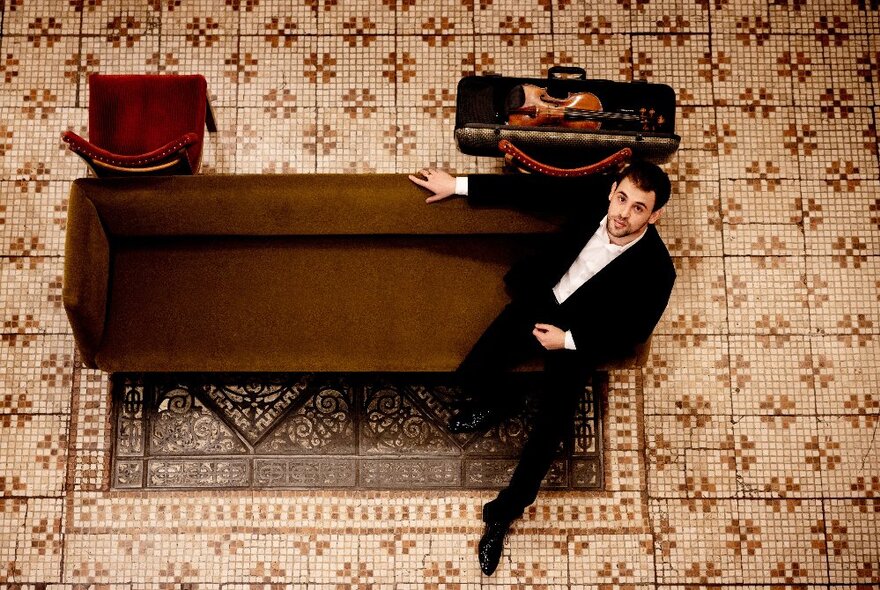 Solo violinist, Yamen Saadi, seated on a brown couch on a brown mosaic-tiled floor, seen from above.