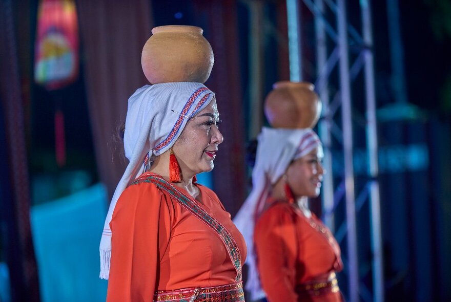 Two women in orange Asian-style traditional dress balancing clay pots on their heads.