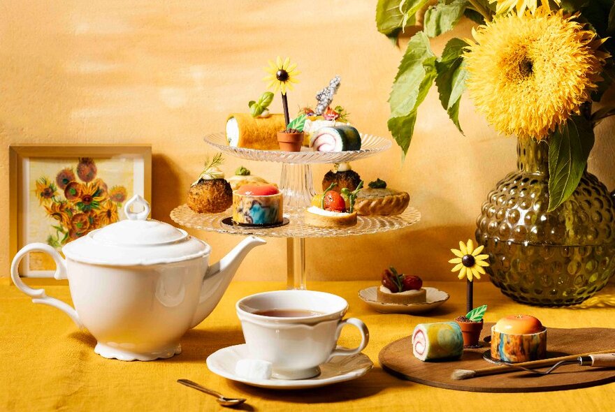 A High Tea setting with a teapot, teacup and saucer, tiered cake tray with small bites and sunflower-themed accessories and a vase with a feathered yellow flower. 