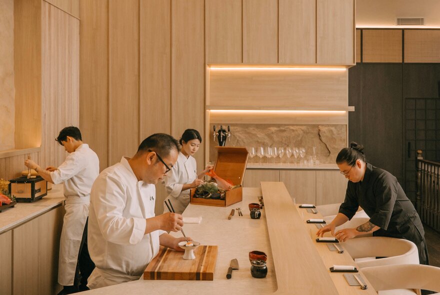 The intimate chef's counter at Sushi Sho, where three Japanese chefs prepare intricate Omakase dishes and a waitress lays place settings at an obvervation bench.