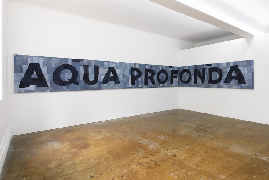 A large sign displayed on a white gallery wall, sewn from recycled denim squares with the words AQUA PROFONDA in black block letters.