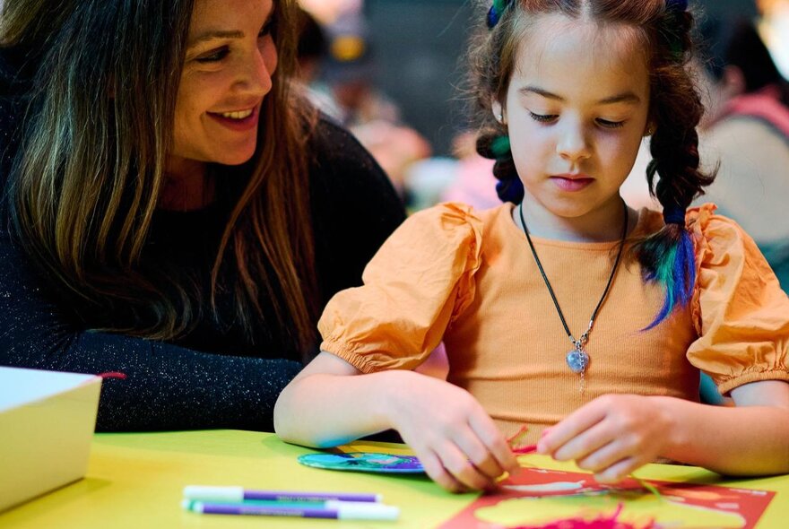 A young child with an adult nearby, taking part in a craft making activity, at the NGV Kids Summer Festival.