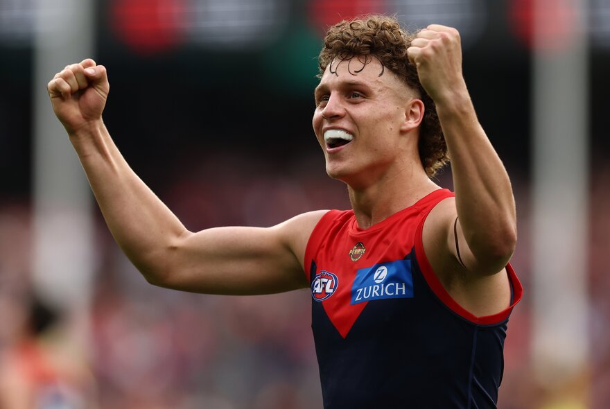 A Melbourne AFL football player on the field during a match with his arms in the air cheering himself on.