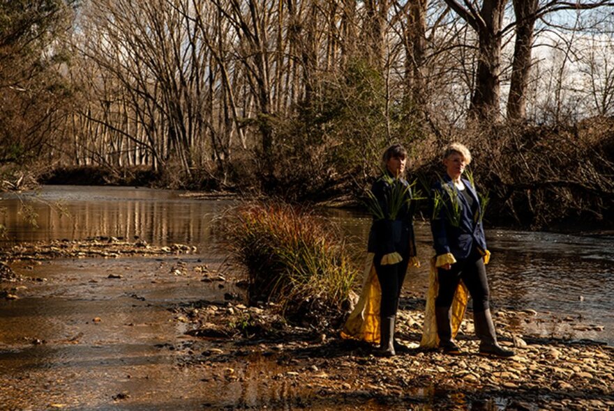 Two people standing on the banks of the Owens River, with leafless trees on the opposite bank behind them.