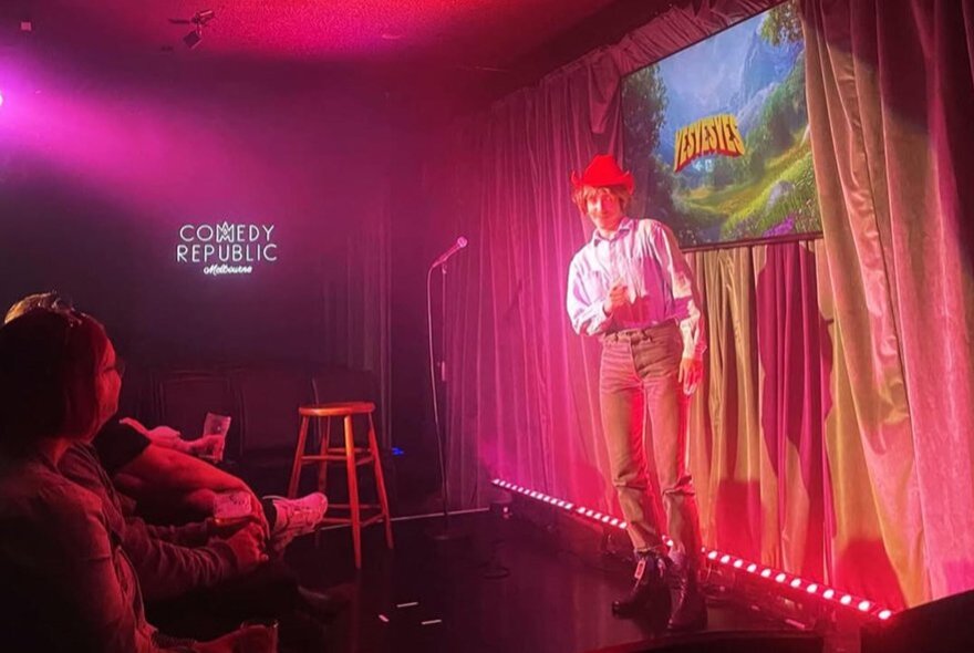 Performer with microphone on small curtained stage in front of seated audience.