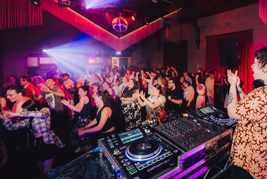 People all doing the same clapping movement on a dancefloor in front of a DJ.