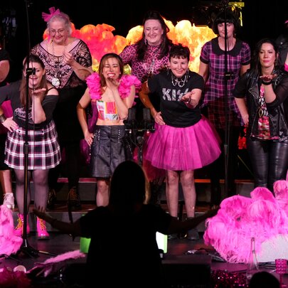 Large choral group wearing pink-themed outfits, standing on a dark stage with pink lights.