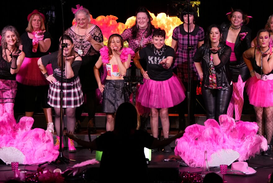 Large choral group wearing pink-themed outfits, standing on a dark stage with pink lights.