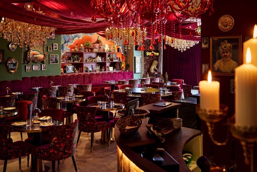 Opulent Indian restaurant interior with tables set for dinner under overhead chandeliers, swags, candles and rich decor.