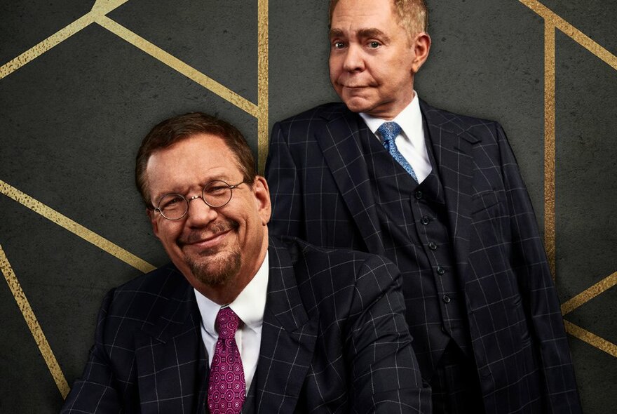 Magicians Penn and Teller, both wearing matching dark suits, smiling at the camera and posed against a dark wall that features gold stripes.