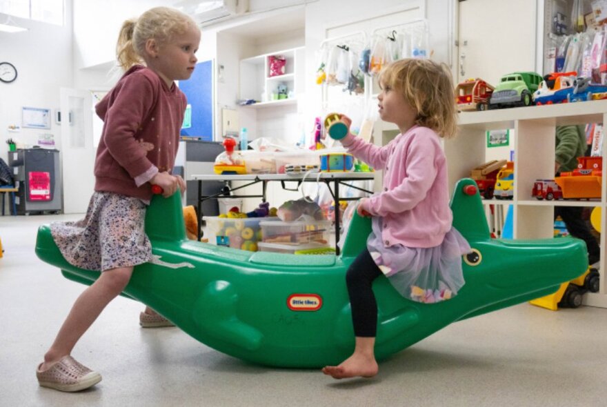 Two young children seated on a green toy crocodile that rocks up and down in a see saw motion, inside a toy library environment.