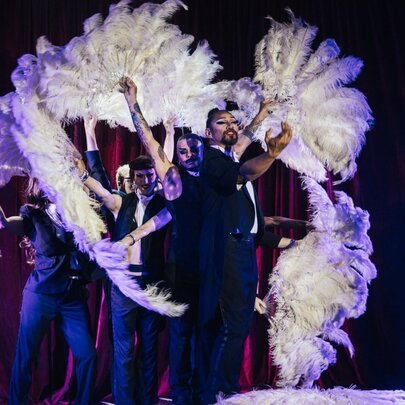 Burlesque performers on stage wearing black clothing and waving white boa fans. 
