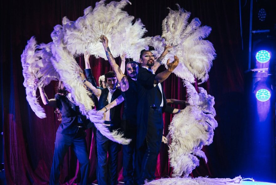 Burlesque performers on stage wearing black clothing and waving white boa fans. 