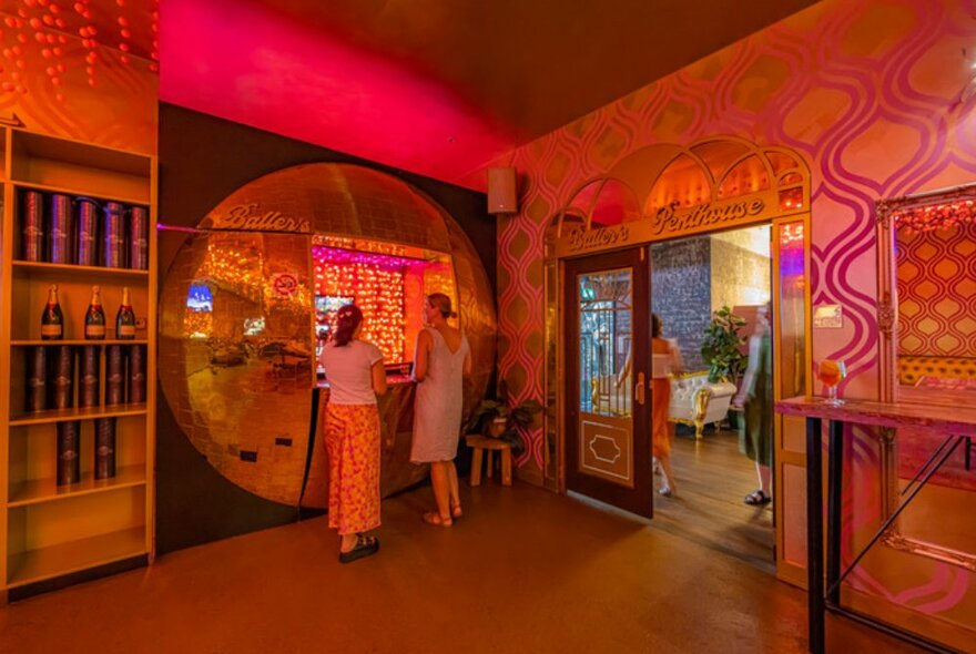 Two girls lining up at the bar, the room is full of neon pink lighting. 