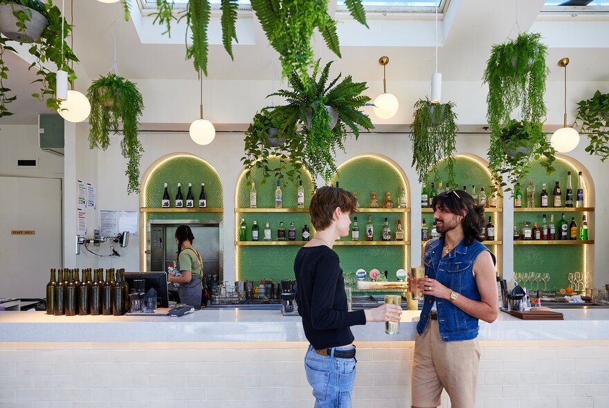 Two men standing at a bar with green tiles and hanging plants.