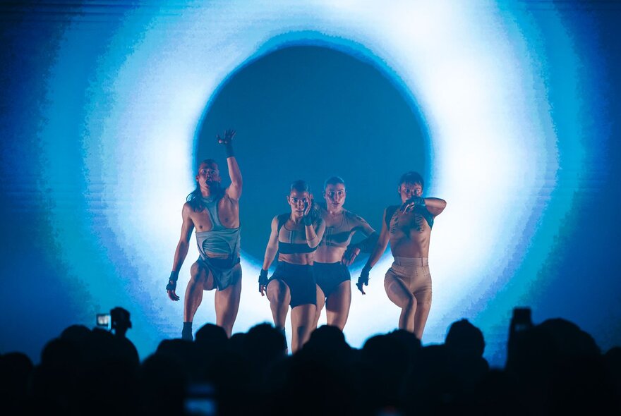 Four performers dancing on a stage in a marching style in front of an audience, with a  blue and white circular backlit screen behind them.