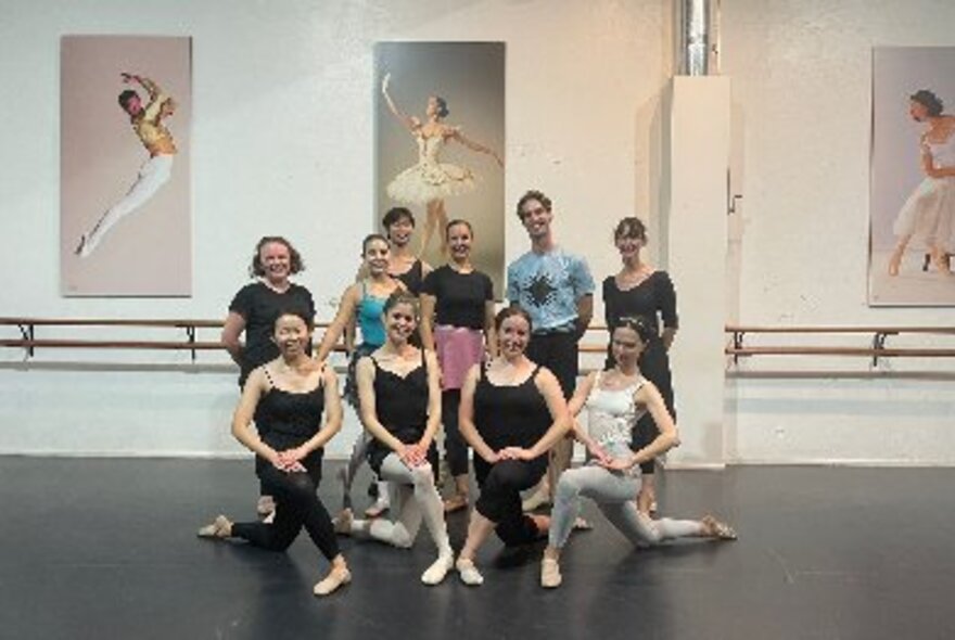 Ballet class participants wearing leotards and leggings, some posing with legs bent, next to a barre in a dance workshop.