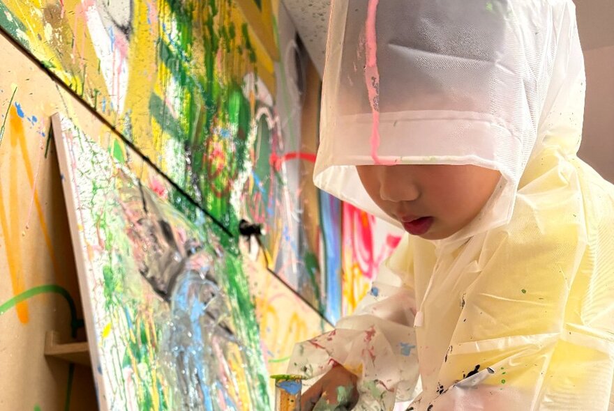 A  child in protective plastic clothing painting onto a canvas.