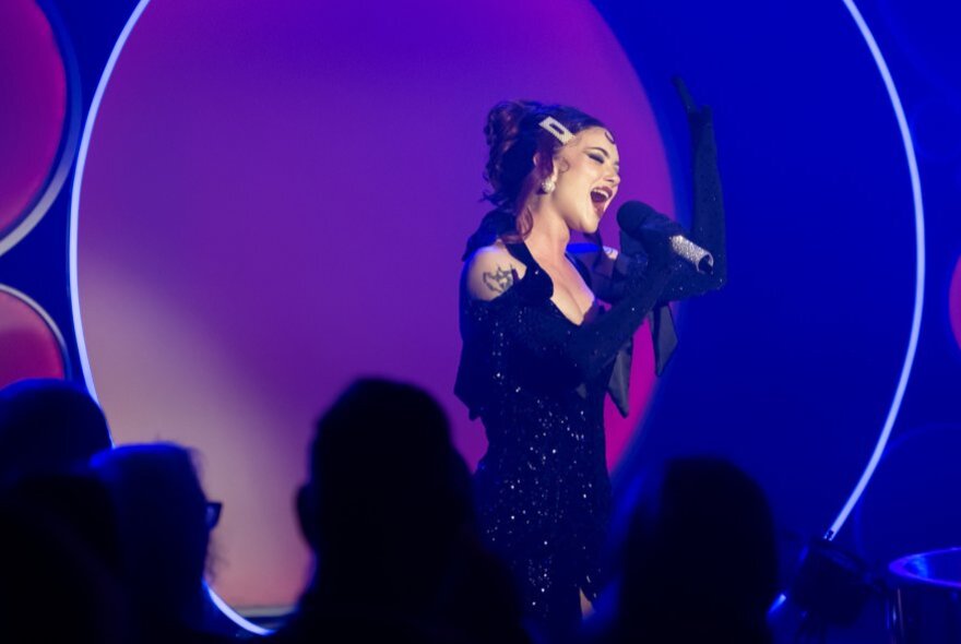 A woman on stage singing into a microphone she holds in her hands, an audience in the dark in front of her.
