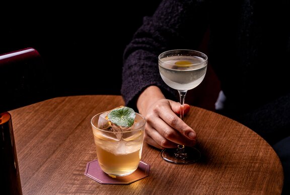 A hand resting on the stem of a cocktail glass served on a small round table, with another cocktail.