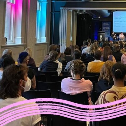A room of seated people watching a panel of speakers with a large video screen, a swish of pink running along the bottom of the image. 