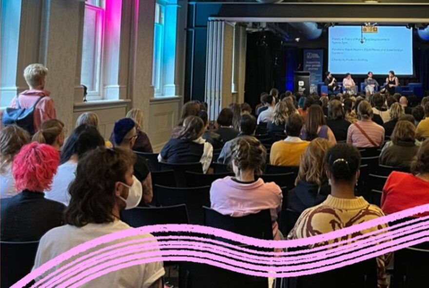 A room of seated people watching a panel of speakers with a large video screen, a swish of pink running along the bottom of the image. 