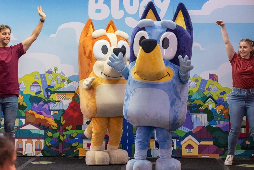 Life size animated dog characters Bluey and Bingo live on stage in an interactive theatre show, with a painted backdrop and two extra human performers dancing alongside.