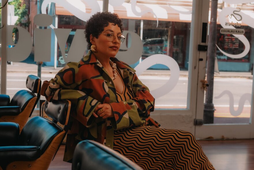 A woman with short, curly hair sitting casually in a shopfront, the street visible through the window behind her. 