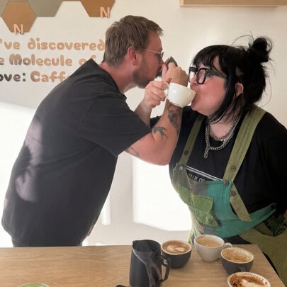 Two individuals interacting closely, drinking from coffee cups, in a barista workshop setting.