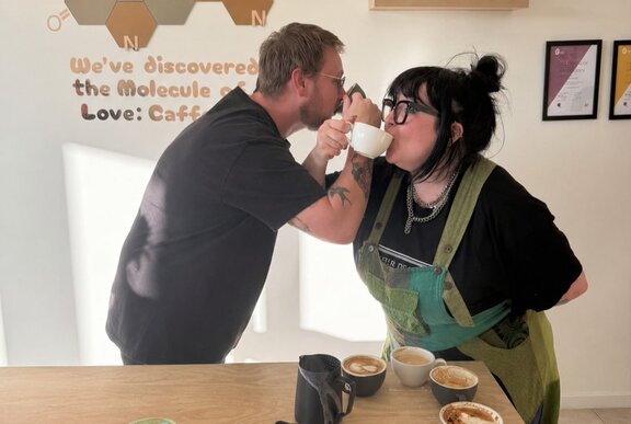 Two individuals interacting closely, drinking from coffee cups, in a barista workshop setting.