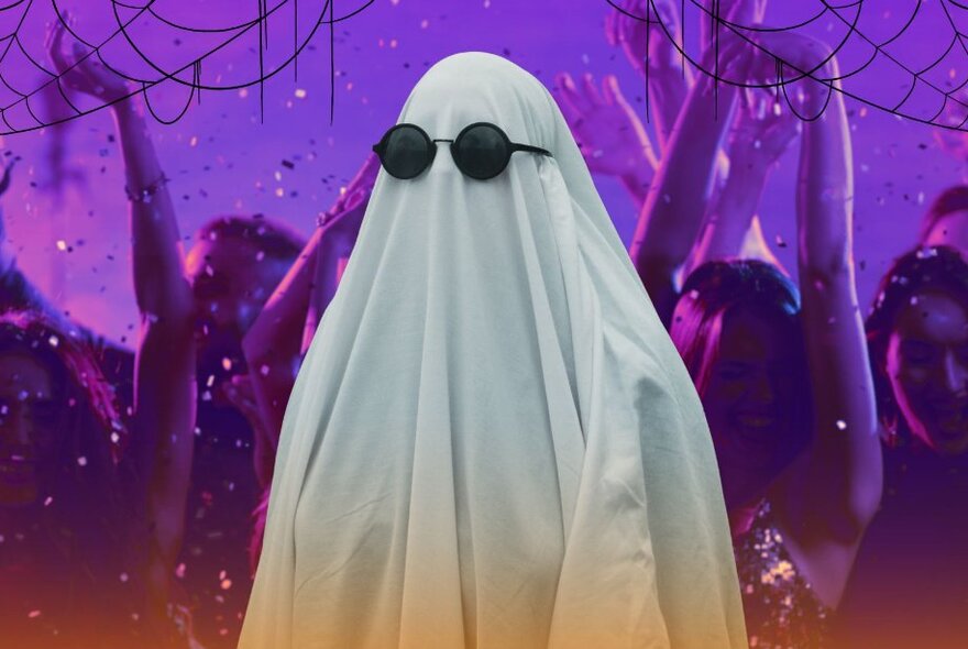 A person dressed as a ghost, wearing a long white sheet over their body, and dark sunglasses on their face, with people dancing in the background.