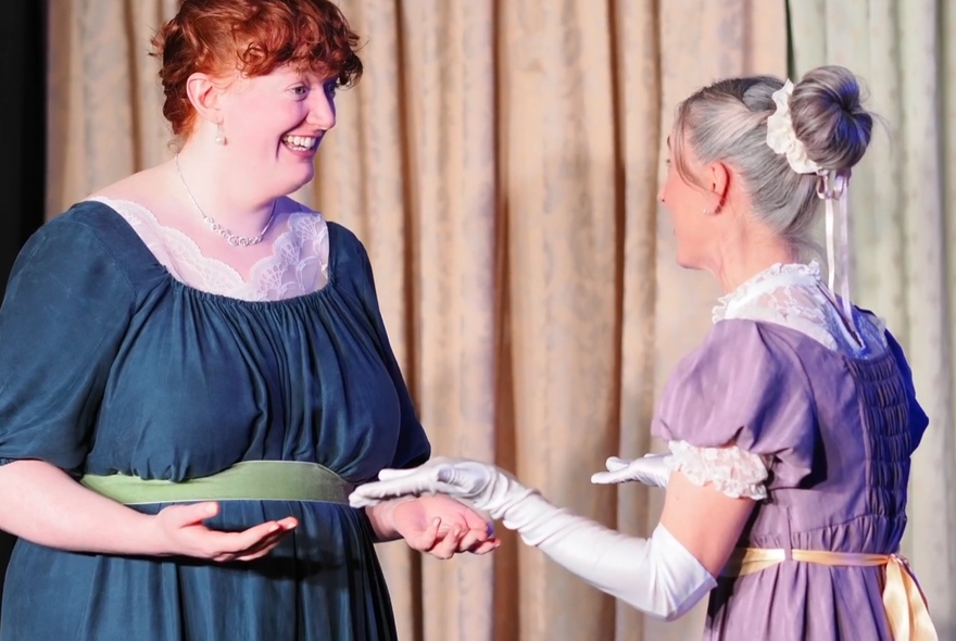Two impro actors on stage in Regency clothing, acting out a scene in front of a curtain.