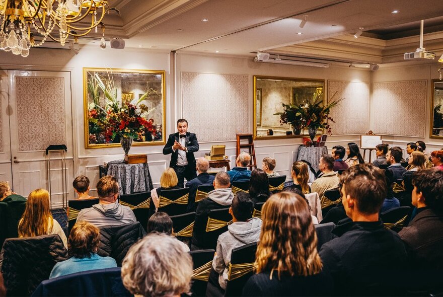 Man wearing a tuxedo and performing magic tricks in front of an audience in a hotel function room. 