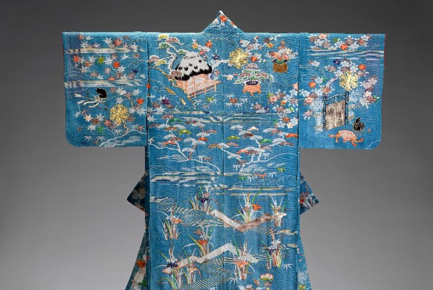 Blue silk kimono with embroidery scenes, gold thread, on display in a gallery.