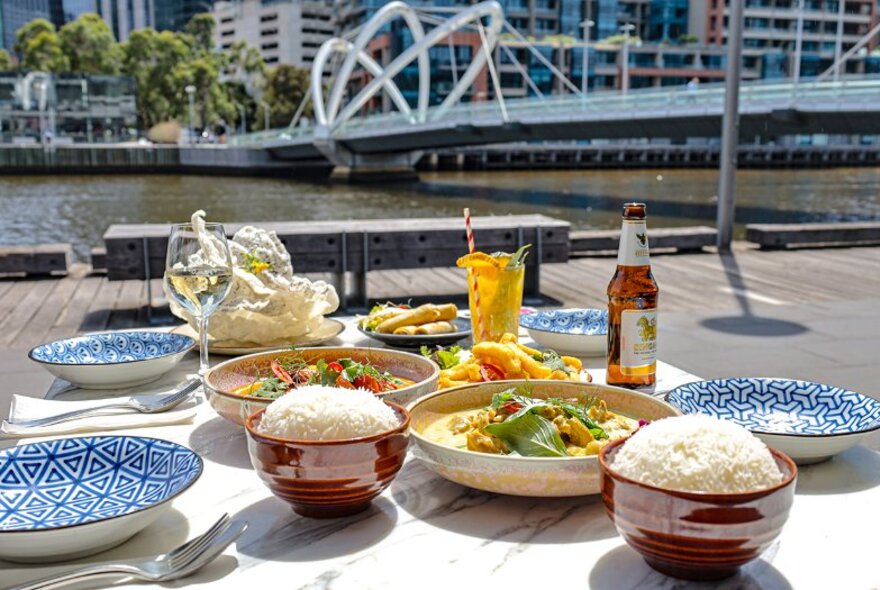 A table with dishes of Thai food, bowls of rice, plates, drinks, and cutlery, set up outside BangPop restaurant on the riverfront, with city buildings in the background.