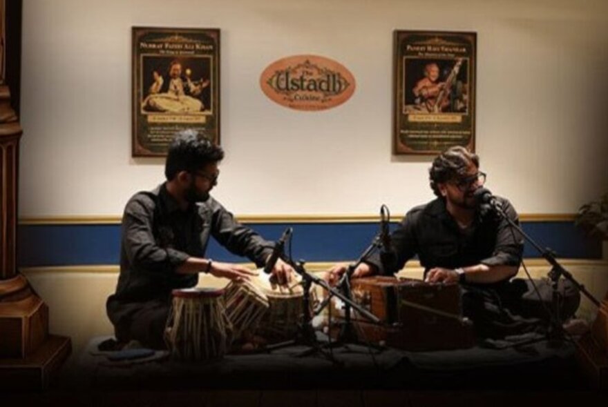 Two seated musicians performing North Indian classical music on a tabla and harmonium inside a restaurant.