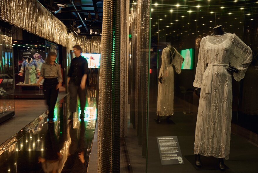 Inside a dimly lit museum two people walk down a corridor on the left while on the right, historical white dresses are displayed on mannequins inside protective glass cases beneath small ceiling spotlights.
