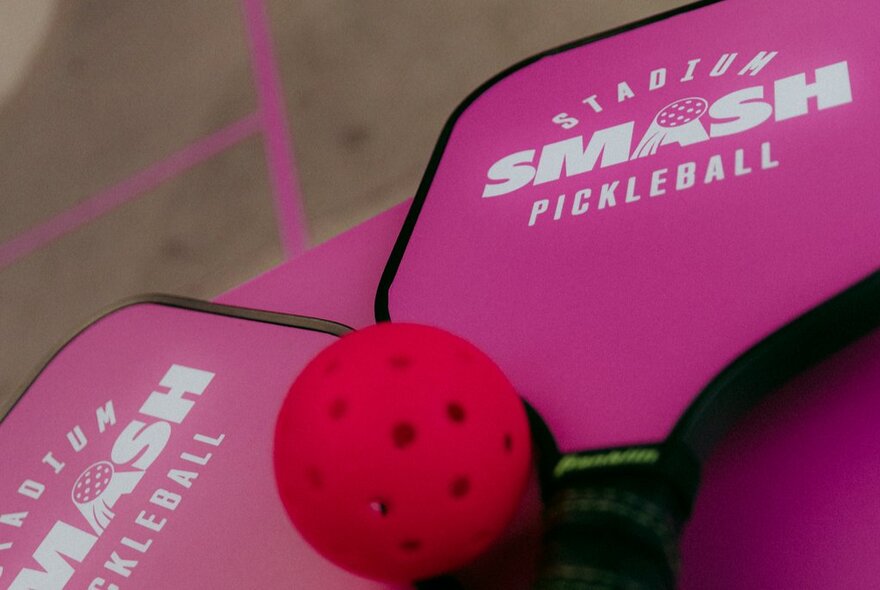 Pink pickleball paddles with white lettering on them that reads Stadium Smash Pickleball, with a small red ball resting on the paddles.