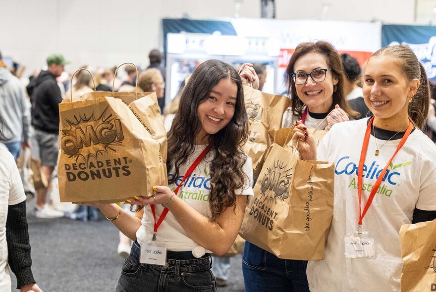 Three smiling people holding up showbags inside a large expo hall.