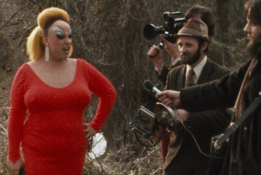 Divine, in character as Edna Turnblad from the film Pink Flamingoes, wearing a bright red dress and a large blonde wig, being interviewed by a man with a microphone, while another person films with a camera.