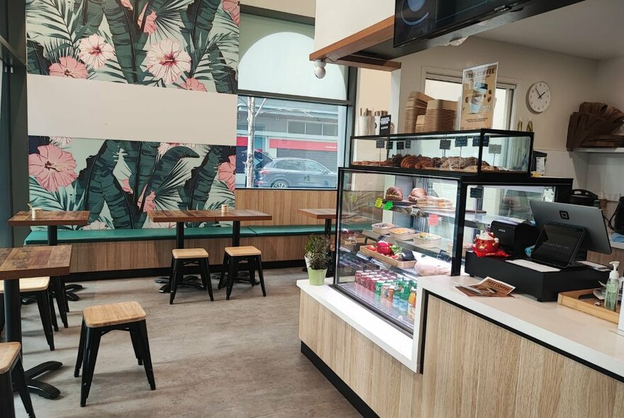 The interior of Merchant 8 cafe, with tables and stools in front of a floral patterned wall and a serving counter with glass display fridge.