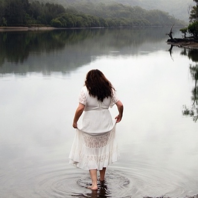 A person with long dark hair wearing a white dress, standing in the shallow water of a calm lake or river surrounded by misty, tree-lined hills.