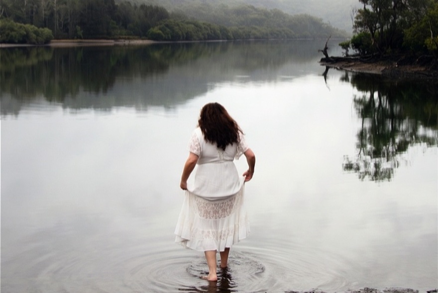 A person with long dark hair wearing a white dress, standing in the shallow water of a calm lake or river surrounded by misty, tree-lined hills.