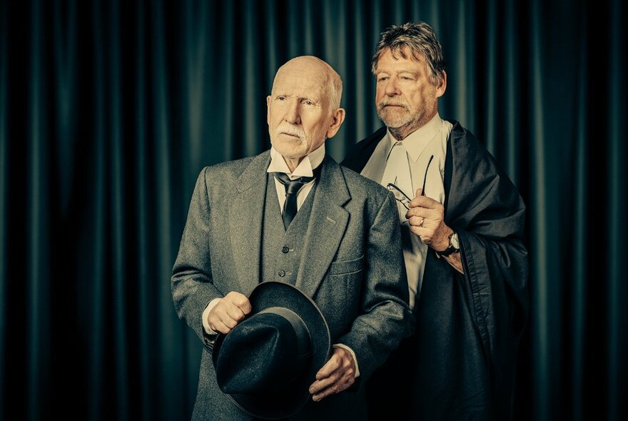 Two actors in costume, standing in front of dark velvet curtains, one wearing a suit and carrying a hat, the other wearing judicial robes.