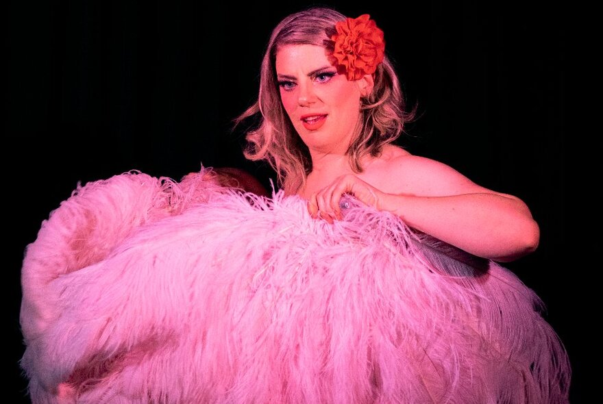 A woman with a flower behind her ear holding up large pink ostrich feather fans. 